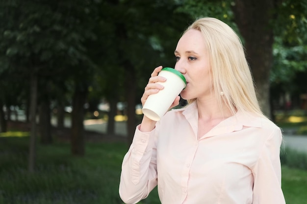 Belle femme d'affaires dans une chemise jupe noire verres boit du café à partir d'une tasse jetable tout en étant assis sur un banc de parc Verre à boisson chaude