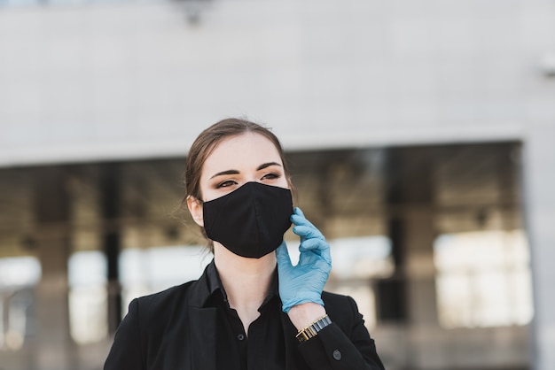 Belle femme d'affaires dans un costume noir dans un masque médical noir et des gants à la ville en quarantaine et isolement. Pandémie COVID-19. Mise au point sélective