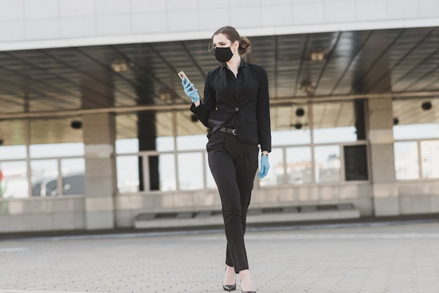 Belle femme d'affaires dans un costume noir dans un masque médical noir et des gants à la ville en quarantaine et isolement. Pandémie COVID-19. Mise au point sélective