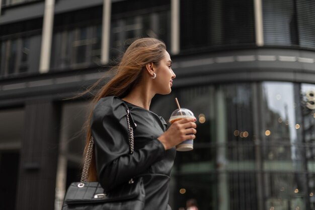 Belle femme d'affaires élégante réussie dans des vêtements noirs à la mode avec un sac en cuir de mode se promène dans la ville et boit du café sur le fond d'un bâtiment moderne de lunettes noires