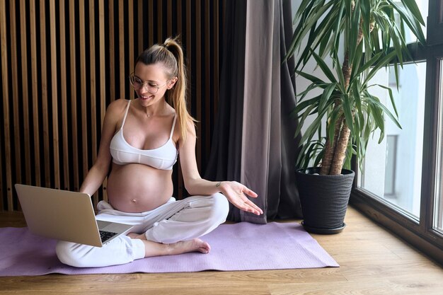 Belle femme d'affaires enceinte à lunettes assise sur le sol sur un tapis à l'aide d'un ordinateur portable et d'un ordinateur portable travaillant à la maison