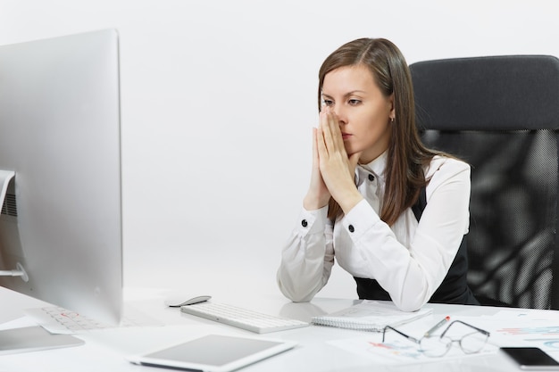 La belle femme d'affaires fatiguée, perplexe et stressée aux cheveux bruns en costume et lunettes assise au bureau, travaillant sur un ordinateur contemporain avec des documents dans un bureau léger