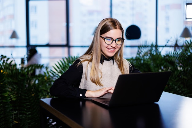 Belle femme d'affaires intelligente et élégante dans une grande entreprise tient une réunion en ligne avec des partenaires commerciaux internationaux, elle est assise à une table sur le lieu de travail avec un ordinateur portable