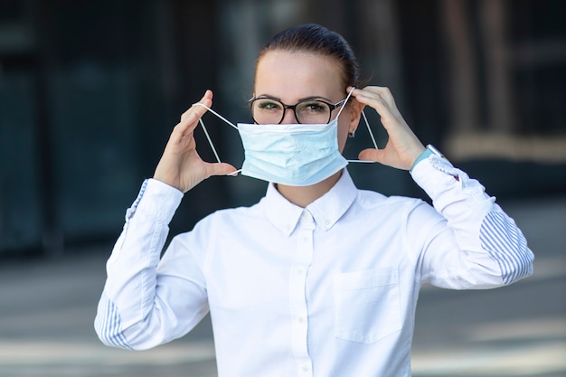 Belle femme d'affaires, jeune fille mettant un masque de protection médical sur son visage, en chemise blanche dans des verres à l'extérieur, soins de santé au travail, travail, bureau. Coronavirus, virus, épidémie, concept Covid-19