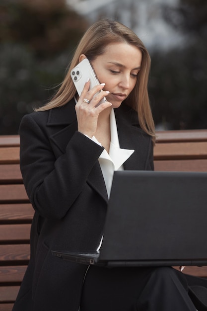 Belle femme d'affaires en manteau noir assis sur un banc avec un ordinateur portable et parler au téléphone