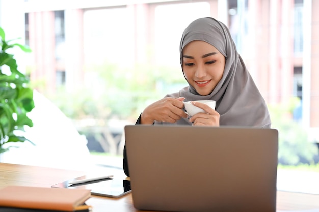 Belle femme d'affaires musulmane avec hijab en sirotant un café du matin en regardant l'écran d'un ordinateur portable