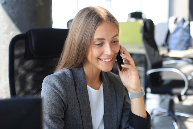 Belle femme d'affaires parle au téléphone mobile et souriant tout en étant assis dans un bureau moderne.