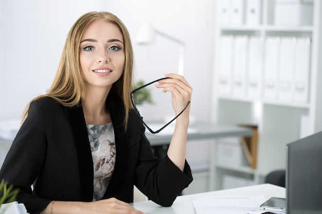 Belle femme d'affaires pensive assise sur son lieu de travail au bureau et tenant des lunettes à la main.