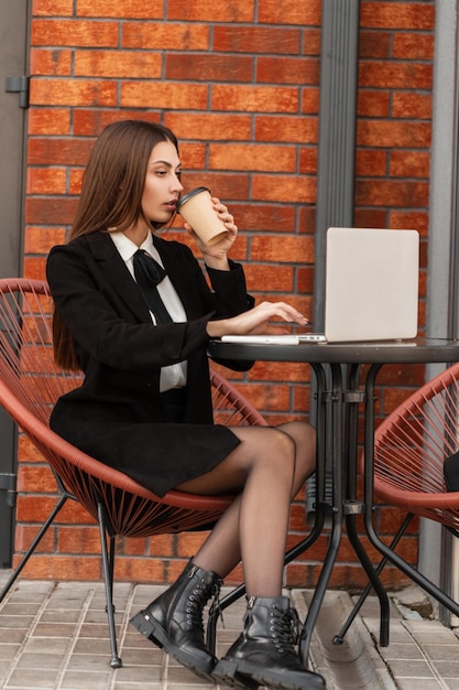 Belle femme d'affaires professionnelle en vêtements noirs de mode avec une jupe et des jambes sexy avec des chaussures est assise et travaille sur son ordinateur portable en buvant du café près du mur de briques