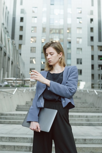 Belle femme d'affaires regardant une tasse de café tenant un ordinateur portable debout dans les escaliers du centre d'affaires