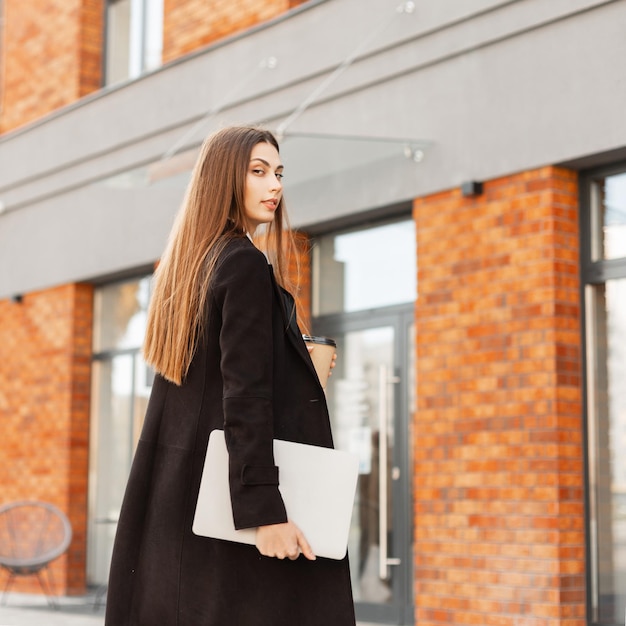 Belle femme d'affaires réussie en tenue de soirée noire avec un café à emporter et un ordinateur portable marchant dans la ville