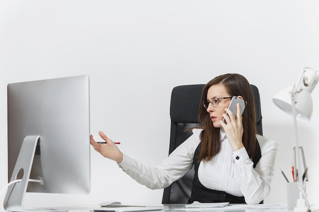 Belle femme d'affaires sérieuse en costume et lunettes assise au bureau, travaillant sur un ordinateur contemporain avec des documents dans un bureau léger, parlant sur un téléphone portable pour résoudre des problèmes,