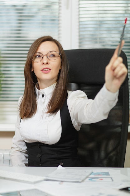 Belle femme d'affaires souriante aux cheveux bruns en costume et lunettes assise au bureau avec tablette, travaillant sur ordinateur avec des documents dans un bureau léger, montrant un crayon de côté