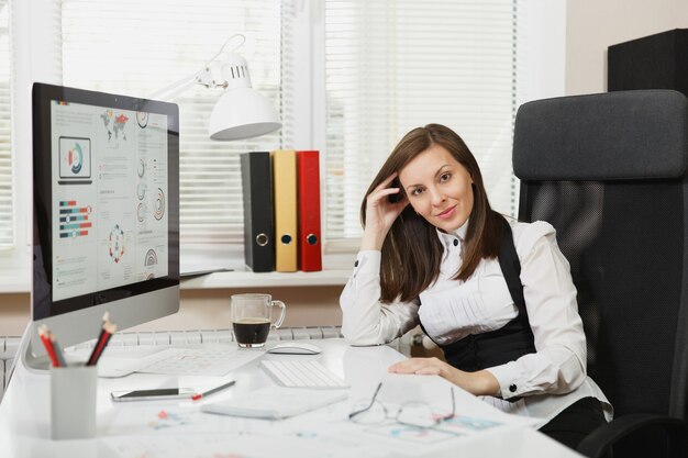 Une belle femme d'affaires souriante aux cheveux bruns en costume et lunettes assise au bureau avec une tasse de café, travaillant sur un ordinateur avec un moniteur moderne avec des documents dans un bureau lumineux, regardant la caméra