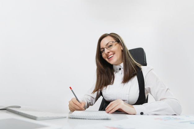 Belle femme d'affaires souriante aux cheveux bruns en costume et lunettes assise au bureau, travaillant à l'ordinateur avec un moniteur moderne et des documents dans un bureau léger, écrivant dans un ordinateur portable