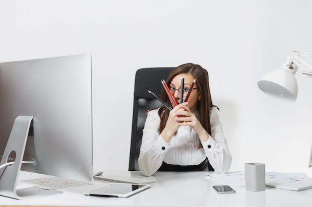 Belle femme d'affaires souriante aux cheveux bruns en costume et lunettes assise au bureau, travaillant à l'ordinateur avec un moniteur moderne avec des documents dans un bureau léger, se cachant derrière des crayons