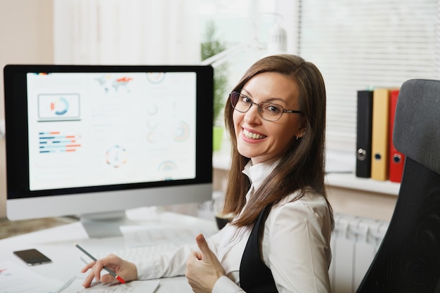 La belle femme d'affaires souriante aux cheveux bruns en costume et lunettes travaillant sur ordinateur avec des documents dans un bureau lumineux, regardant la caméra, contre le mur