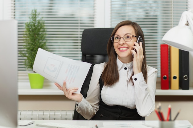 Belle femme d'affaires souriante en costume et lunettes assise au bureau, travaillant sur un ordinateur contemporain avec des documents dans un bureau léger, parlant au téléphone portable, menant une conversation agréable