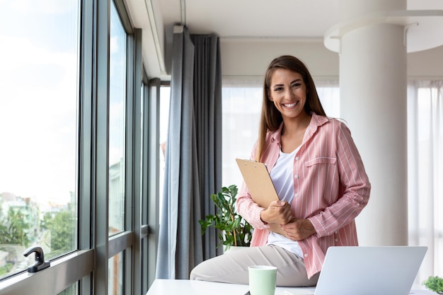 Belle Femme D'affaires Souriante Lisant Quelque Chose Sur Un Ordinateur Portable Tout En S'appuyant Sur La Table Du Bureau