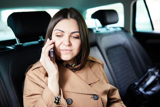 Belle femme d'affaires utilise un téléphone intelligent et sourit assis sur le siège arrière de la voiture