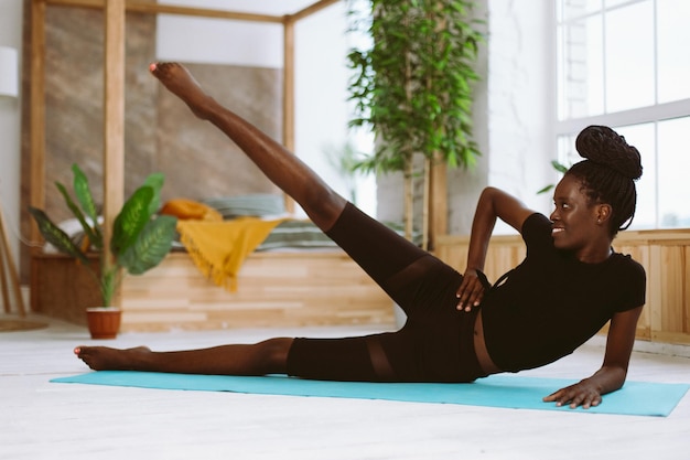 Belle femme afro-américaine athlétique allongée sur le côté et levant la jambe vers le haut formation de mise au point sélective sur un tapis de gym dans un studio photo décoré Cours de sport technique d'exercice fitness garder la forme du corps