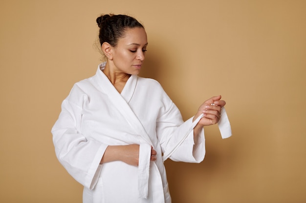 Belle femme afro-américaine attachant une ceinture sur une robe gaufrée blanche posant contre une surface beige avec espace de copie. Concept de soins de la peau et de spa