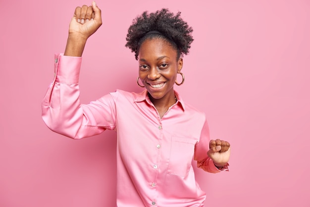 Belle femme afro-américaine aux cheveux bouclés danse avec joie garde les bras levés étant de bonne humeur se sent très heureux