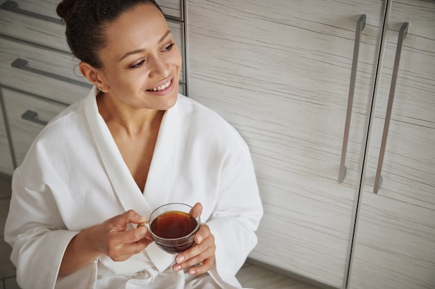 Belle femme afro-américaine avec un beau sourire à pleines dents en peignoir blanc regardant loin avec une tasse de café dans les mains.