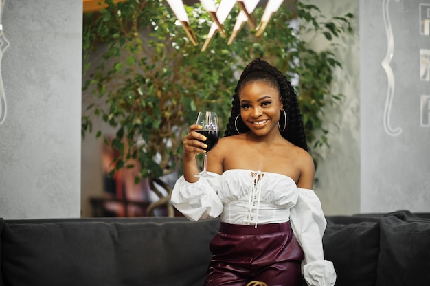 Belle femme afro-américaine en chemisier blanc et pantalon en cuir rouge pose au restaurant avec un verre de vin.