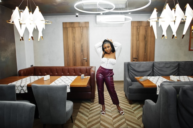 Belle femme afro-américaine en chemisier blanc et pantalon en cuir rouge pose au restaurant.