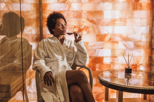 Belle femme afro-américaine mûre buvant du vin rouge et se relaxant dans un sauna de grotte de cristal de sel au centre de spa.