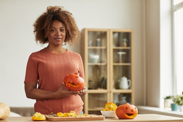 Belle femme afro-américaine portant une tenue décontractée debout à la table de cuisine tenant la citrouille sculptée pour Halloween