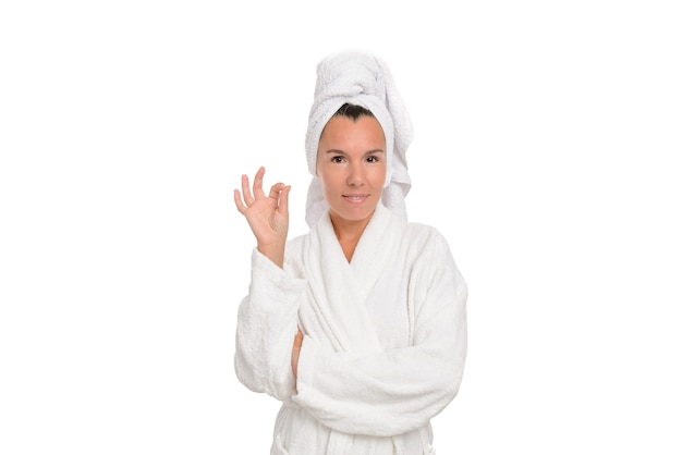 Une belle femme d'âge moyen dans une robe blanche et une serviette sur la tête sourit et montre un geste OK