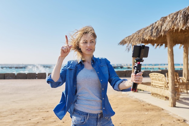 Belle femme d'âge moyen enregistrant une vidéo sur fond de plage mer coucher de soleil smartphone