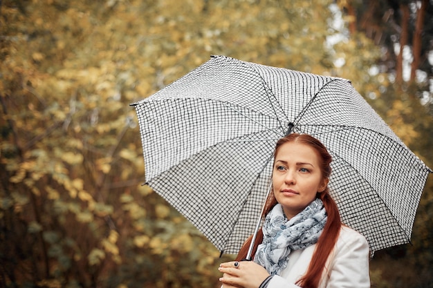 Belle femme d'âge moyen de race blanche aux cheveux rouges avec un parapluie dans le parc par une journée d'automne nuageuse.