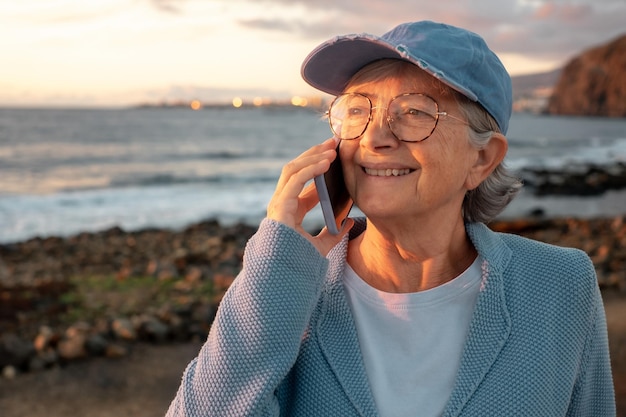 Belle femme âgée parlant au téléphone debout sur la plage de la mer au lever du soleil