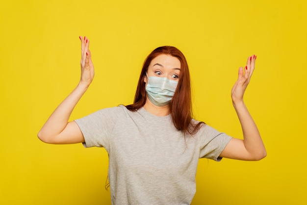 Une belle femme agite ses bras et est choquée par la propagation du coronavirus. Belle fille rousse dans un masque de protection sur un mur jaune. Le concept de protection et de prévention