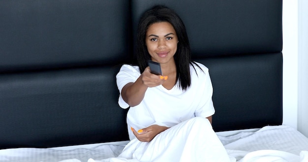 Belle femme allongée sur un lit avec télécommande et regarder la télévision.