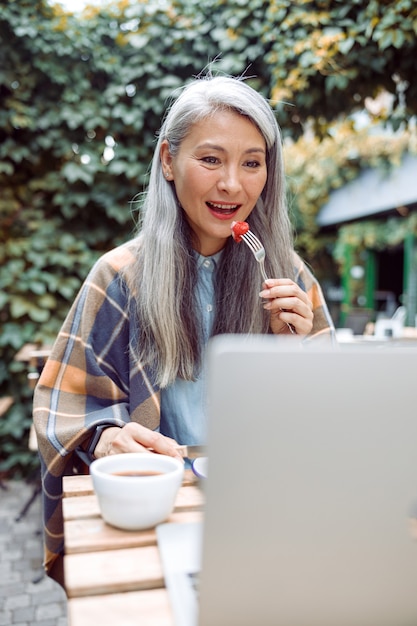 Belle femme asiatique âgée mange des fraises fraîches devant un ordinateur portable à table à l'extérieur