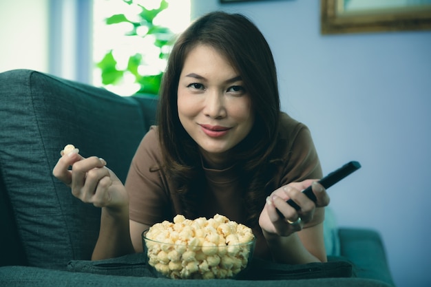 Belle femme asiatique allongée sur un canapé pour se détendre et tenir la télécommande à la main avec un bol de pop-corn dans le salon de la maison.