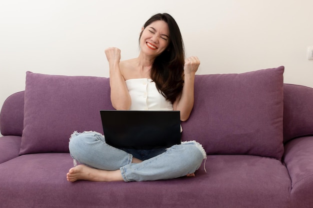 Une belle femme asiatique assise sur un canapé avec un ordinateur portable lève le bras avec succès ou célèbre le shopping en ligne