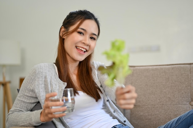 Belle femme asiatique assise sur un canapé tenant un verre d'eau et de laitue verte