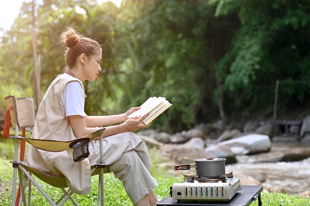 Belle femme asiatique assise sur la chaise de camping se détendre en lisant un livre près de la rivière