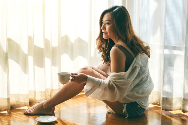 Une belle femme asiatique assise et tenant une tasse de café chaud à boire par terre le matin