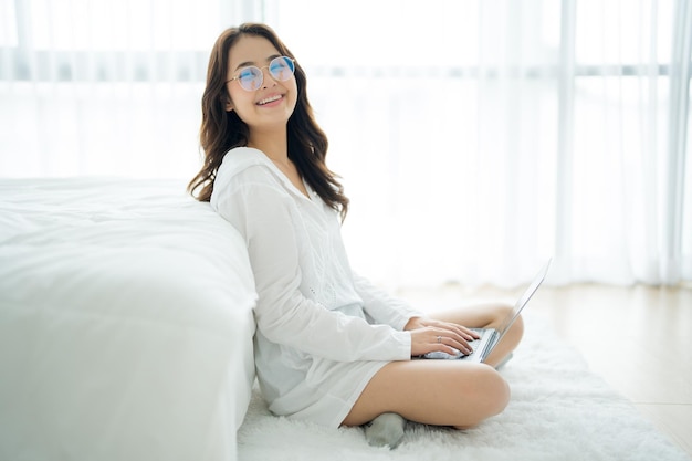 Une belle femme asiatique aux cheveux longs est assise près du lit et utilise son ordinateur portable