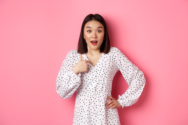 Belle femme asiatique brune pointant le doigt vers la caméra, semblant surprise d'être choisie, debout en robe sur fond rose.