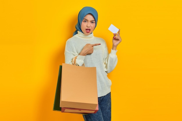 Belle femme asiatique choquée en chandail blanc tenant un sac à provisions et pointant du doigt la carte de crédit isolée sur fond jaune