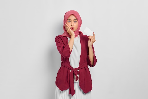 Belle femme asiatique choquée en chemise décontractée et hijab touchant la joue du visage, tenant une carte de crédit avec la bouche ouverte isolée sur fond blanc. Concept de mode de vie religieux de personnes