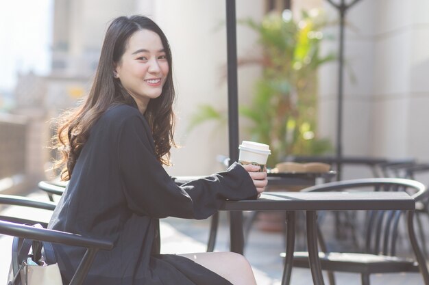 Une belle femme asiatique en costume bleu foncé est assise sur une chaise tenant une tasse de café