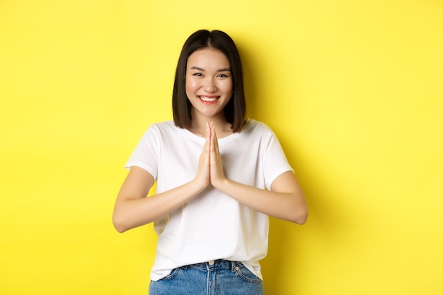 Belle femme asiatique dire merci, se tenant la main en namaste, prier le geste et souriant, être reconnaissante, debout sur fond jaune.